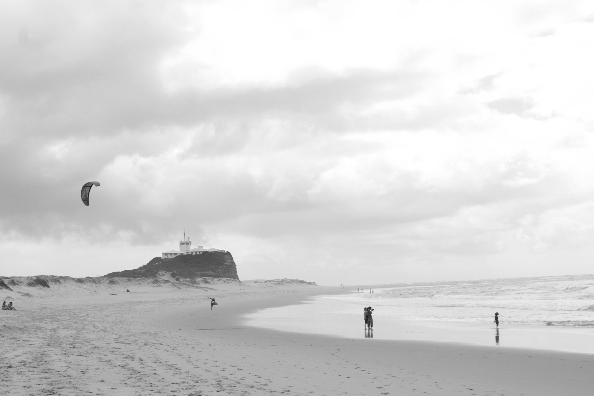 People and a kite appearing small along a broad beach beneath a cliff and expansive sky